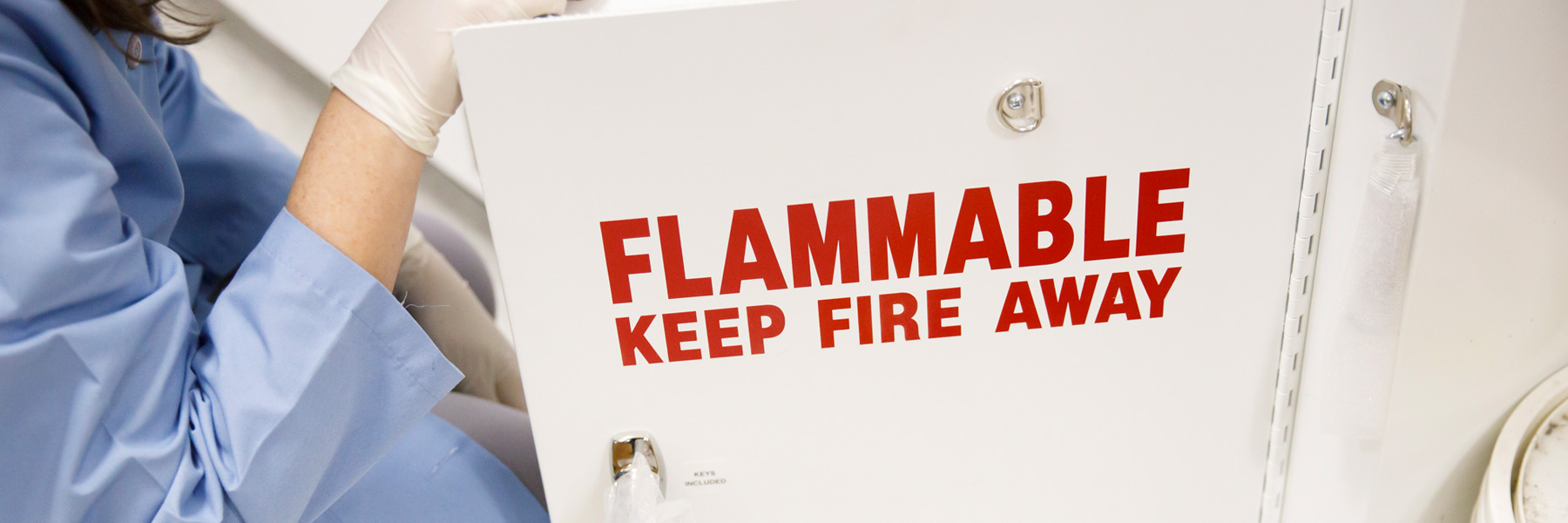 A woman in a lab coat and protective gloves opens a metal cabinet labelled FLAMMABLE KEEP AWAY in bright red letters.