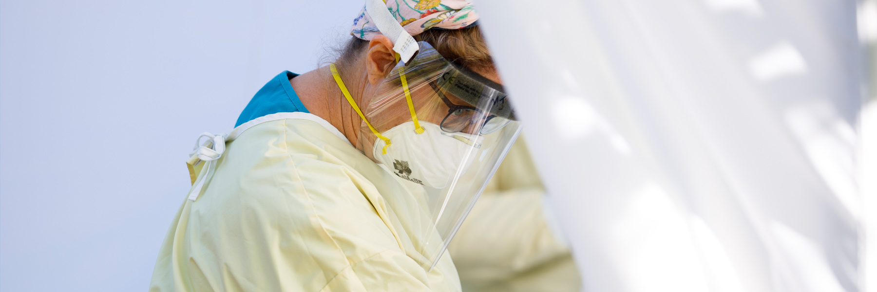 A bright, airy photo of a nurse wearing PPE working in a tent. Her soft yellow scrubs and the white tent background draw the point of focus to her worried expression.