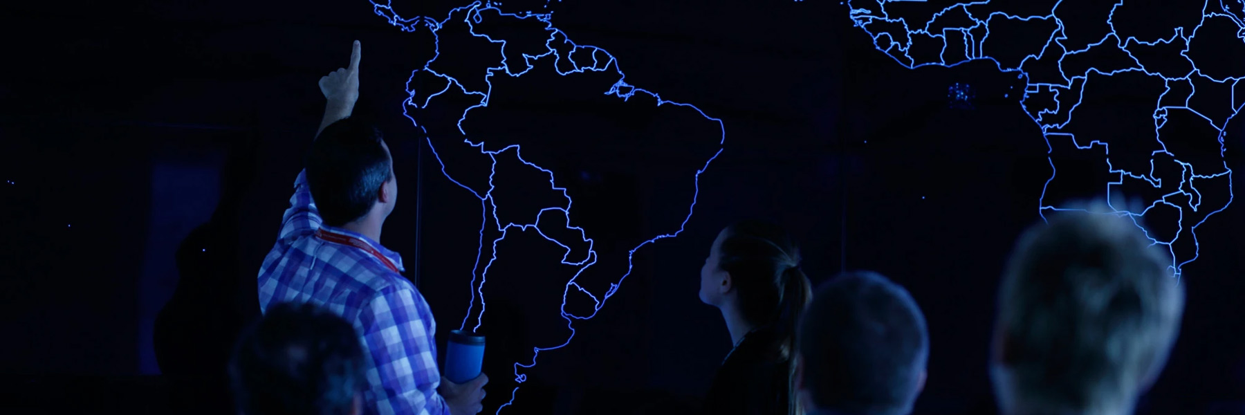 A dramatic darkly lit photograph of a man pointing to a spot on a world map in front of a crowd. 