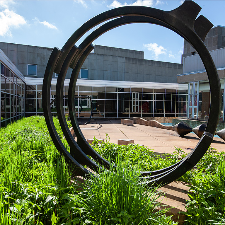 A circular sculpture amidst greenery frames the front of an IU Northwest campus building.