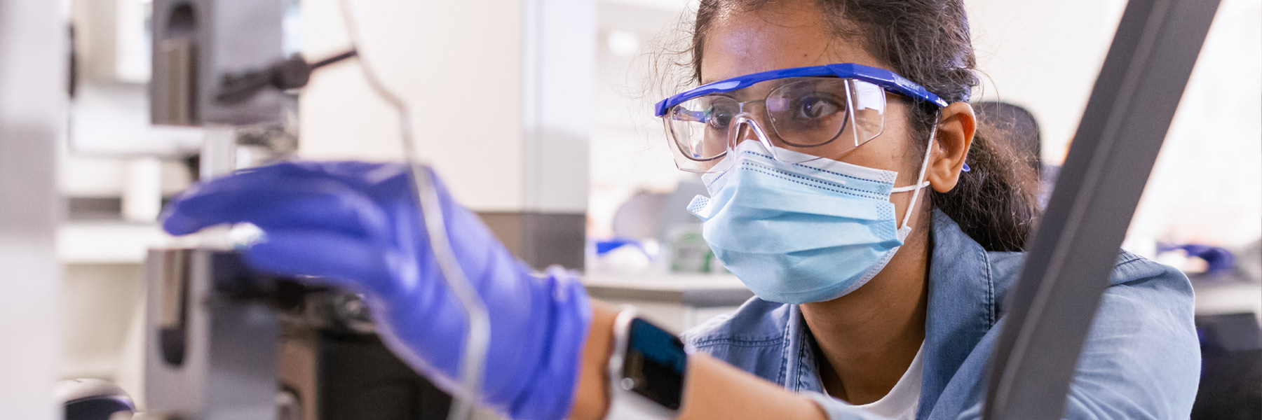 A young woman in safety glasses, mask, and gloves examines a piece of biomedical equipment.