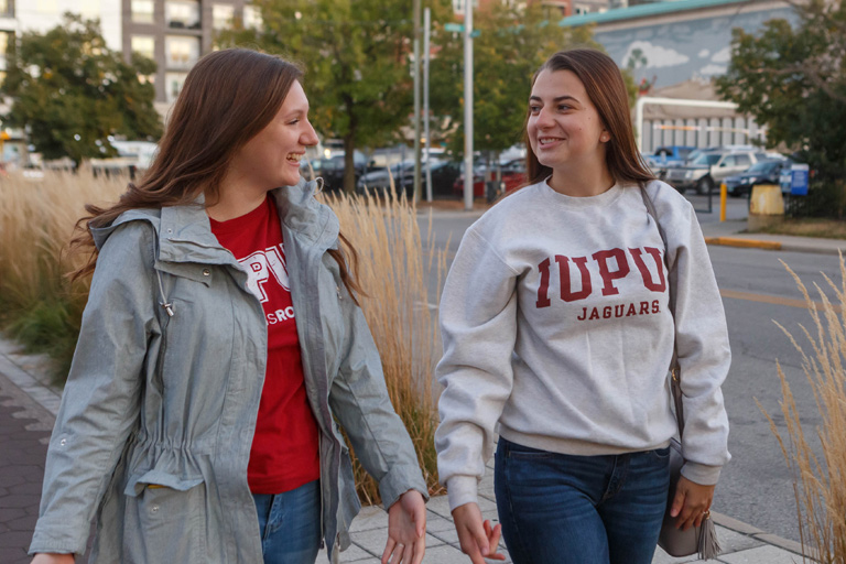 Two young women wearing IUPUI gear take a walk together down the Indianapolis Cultural Trail at dusk. 