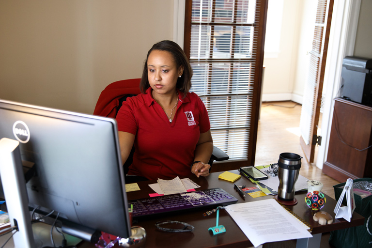 A distinguished-looking faculty advisor in business wear sits comfortably at her desk, piled with papers, as she speaks with a student in an airy office. 