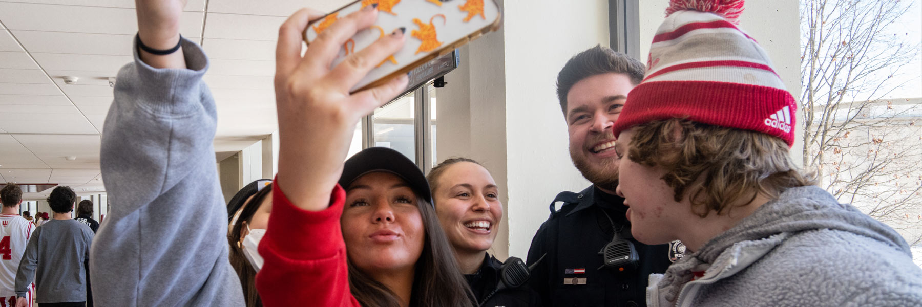 Two young adult women with a tabby cat cell phone case wearing Indiana gear take a selfie with a grinning IUPD cadet indoors.