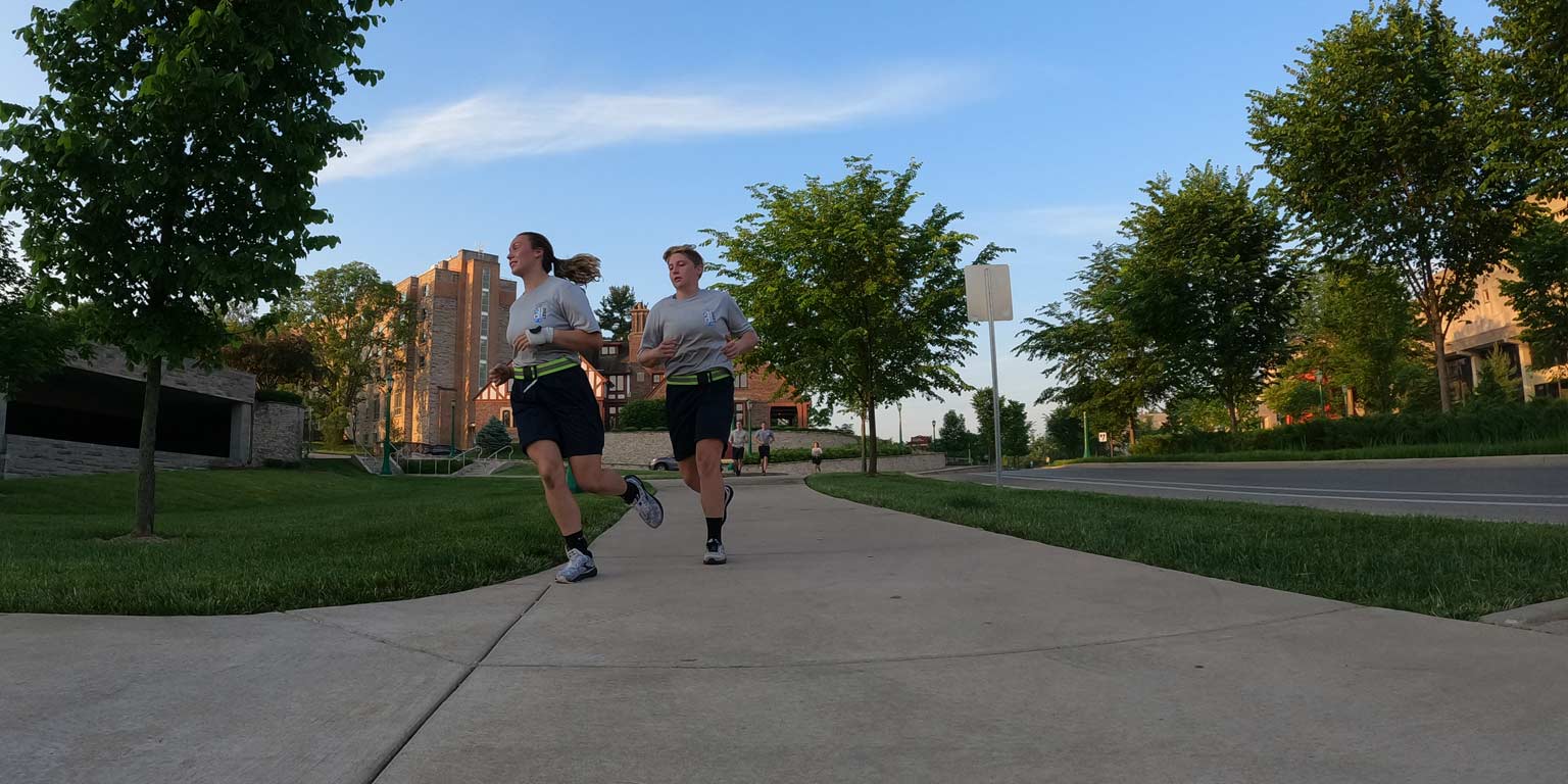 Two IU Police Academy cadettes in physical exercise uniforms run down Eagleson Avenue in the cool of a summer evening.