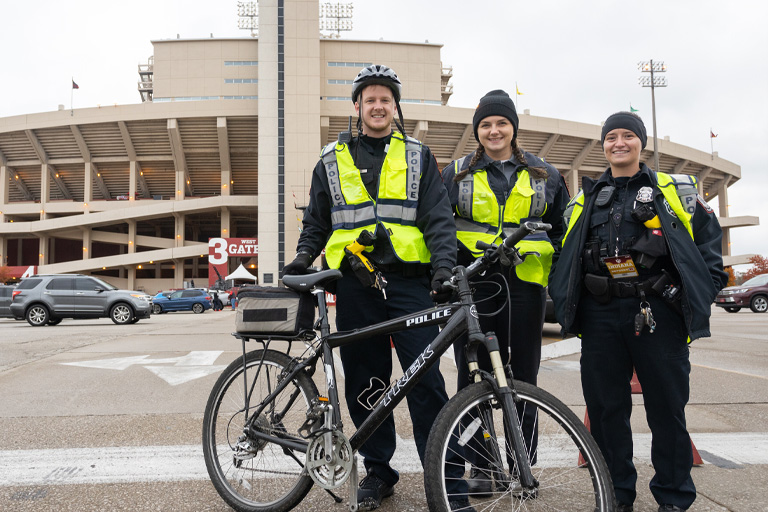 An IUPD bicycle officer grins with two colleagues outside Memorial Stadium, on a chilly fall day. They are wearing winter uniforms, with high-visibility vests and earwarmers.