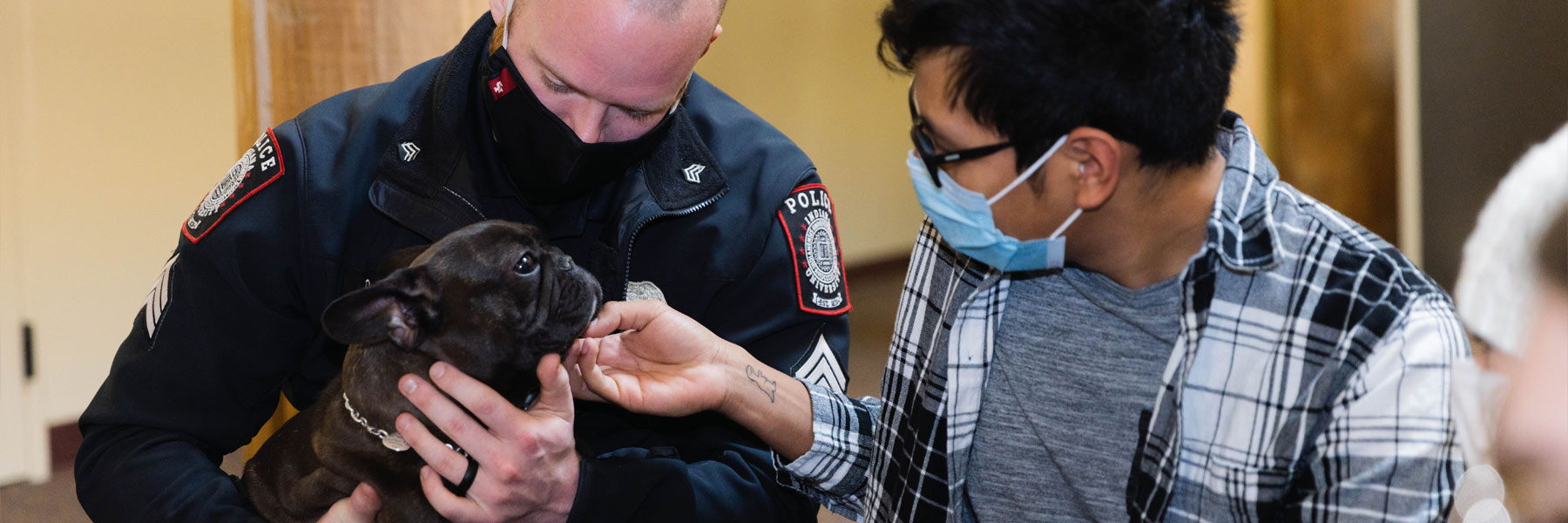 A young masked student tickles the chin of a French Bulldog being held by a member of IU Southeast's Police department during a mental health support animal event.