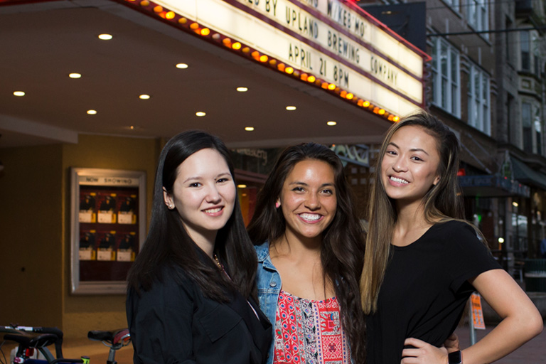 Three young women out for a night on the town pose in front of a brightly lit theatre marquee. 