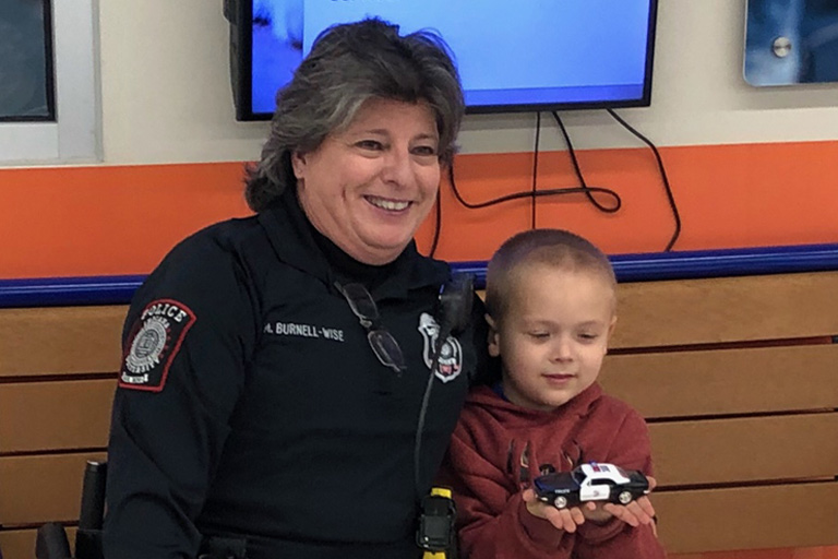 A grey-haired female IUPD officer smiles for a photo with a little boy holding a toy police car.