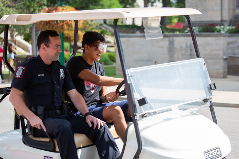 A student wearing drunk goggles attempts to operate a golf cart under the supervision of an IUPD officer as part of a driving impairment demonstration. The officer, sitting beside the student, is smiling but tightly gripping the vehicle's armrest.