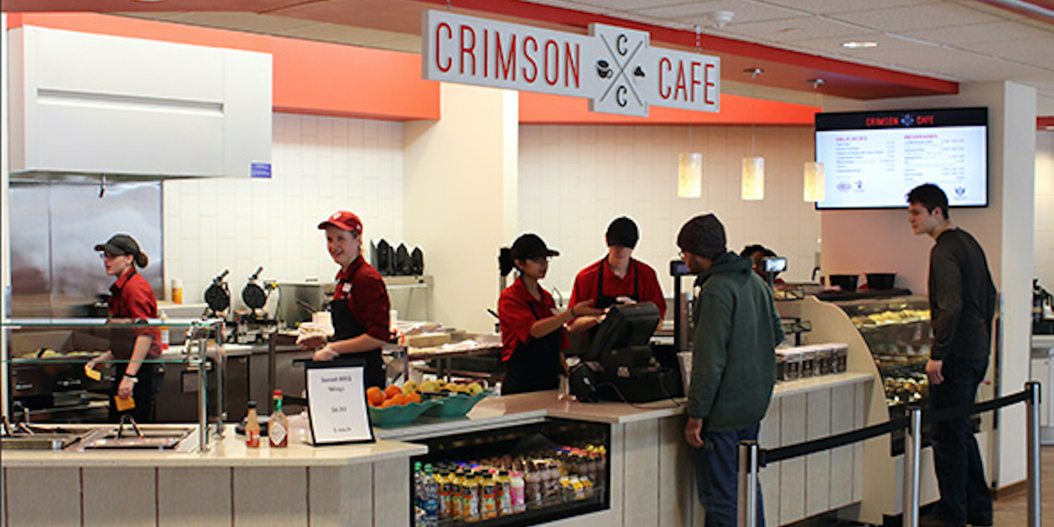 Four people working behind the counter under a sign that says "Crimson Cafe" and  two customers